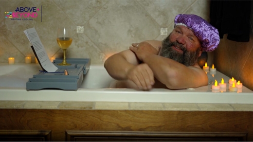 A man sits in a bubble bath wearing a purple shower cap.